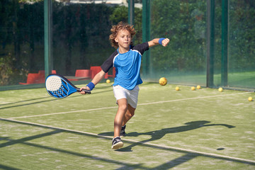 Full body concentrated boy in sportswear swinging paddle while playing paddle tennis on sunny day on court