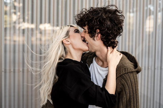 Side view of cheerful adult blond woman biting for tongue and touching neck of young curly dark haired man while standing and kissing with pleasure on street against striped wall of modern building in windy weather
