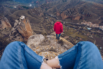 Crop from behind person in jeans sitting with crossed legs while man in jacket gazing and standing on top on mountain