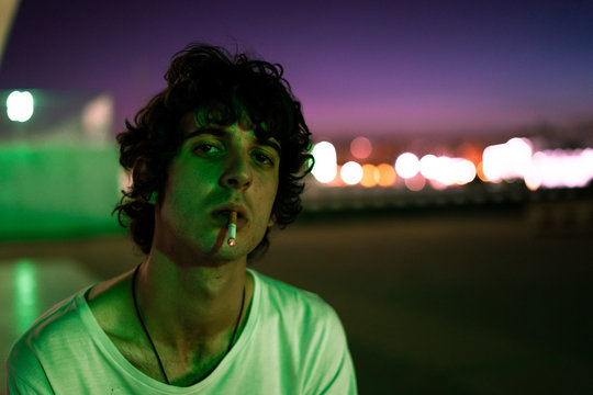 Handsome provocative confident guy in casual clothes with cigarette in mouth looking with interest at camera against blurred background of street under purple sky during sunrise