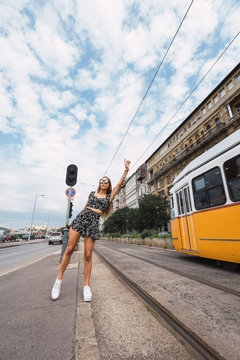 Active slim woman raising hand waiting tram nearby railway in bright cloudy day in Budapest