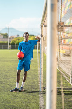 Full body African American teenager in sportswear holding ball and leaning on net during training on football field