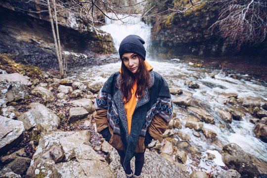 Woman in warm jacket standing by furious torrent at evergreen forest on cold autumn day and looking at camera