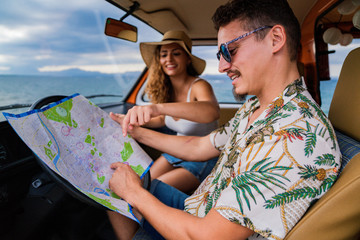 Side view of active smiling man in sunglasses looking at open road map and finding route with woman in hat in car