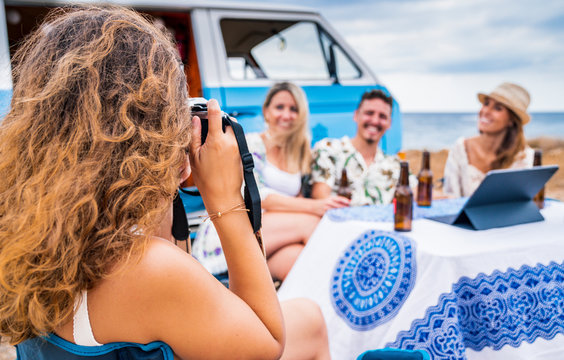 Cheerful friend smiling and holding bottle of drinks sitting at table while long haired woman taking photo nearby blue car