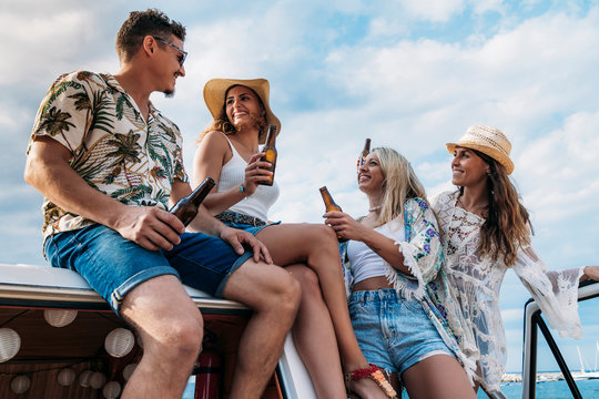 Cheerful lazy ladies with long hair in hat raising bottles of drink and clinking with stylish man in car roof