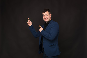 Cheerful young man in blue suit pointing to copy space on a black background.