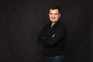 Fototapeta premium Smiling man in black shirt is sitting with crossed hands and looking at the camera near black wall.