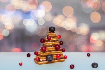 Homemade Christmas tree shape honey biscuit cake with berries on white kitchen table winter window background with bokeh lights city view