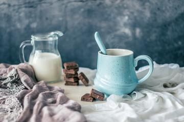 Blue pastel coffee mug, bottle of milk and chocolate on a rustic white table and fabric