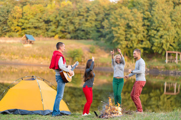 Group of happy friends with guitar, having fun outdoor, dancing and jumping near the lake in the park background the Beautiful sky. Camping fun