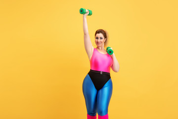 Young caucasian plus size female model's training on yellow background. Copyspace. Concept of sport, healthy lifestyle, body positive, fashion, style. Stylish woman practicing with weights and smiling
