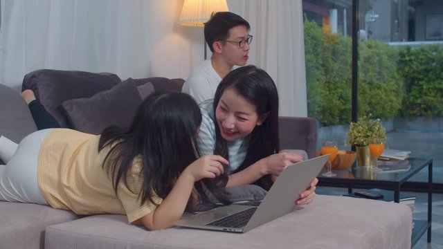 Asian family enjoy their free time relax together at home. Lifestyle mom and daughter using laptop watch movie on internet, dad watch TV in living room in modern home concept.