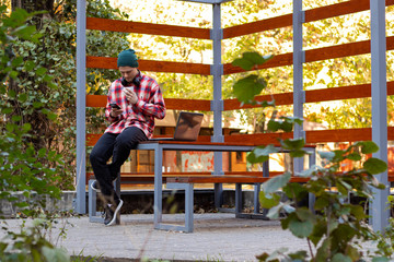 Stylish male freelancer working on new project, drinking coffee and waiting for video call while analyzing data using laptop computer and wireless connection to 4G internet in city parks gazebo. 