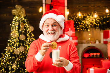 Happy Santa Claus eating a cookie and drinking glass of milk at home Christmas interior. Santa Claus enjoys cookies and milk left out for him on Christmas eve.