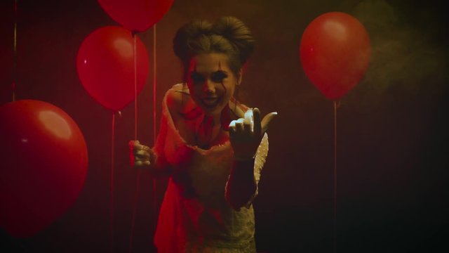 insidious young woman in image of crazy clown waves her hand calls for lures to go with her in her hand is red ball. Concept of childhood fear and phobia. Creative hairstyle professional art makeup.