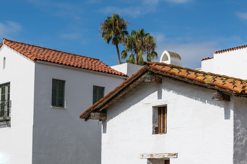 Old and new Spanish style buildings with white plaster walls and red tile roofs with palm trees under a blue sky in Santa Barbara, California, USA