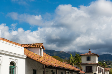 Red tile roof and white Spanish style buildings beneath palm trees, mountains, and dramatic white fluffy clouds in Santa Barbara, California - obrazy, fototapety, plakaty