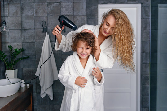 Beautiful mom dries hair with a hairdryer to her little happy son, dressed in a white bathrobe.