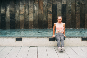 Outdoor portrait of young beautiful fit woman doing tricep dips, wearing fashionable activewear, athlete model posing in urban background, sport fashion