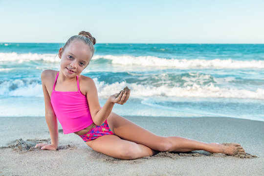 A playful cute little girl in a colorful pink swimsuit sitting on the beach, waves, holding sand in hand, ocean background, copy space