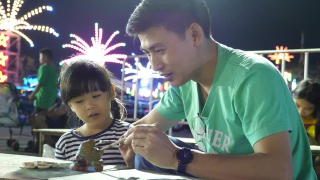 Asian father and daughter painting plaster crab toy together at night festival, family bonding activity
