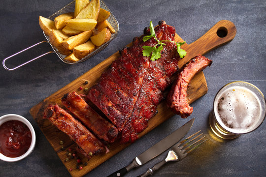Pork loin ribs served on chopping board, potato wedges and glass of beer. View from above, top