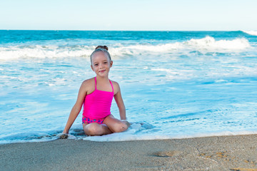 Fototapeta na wymiar A playful cute little girl in a colorful pink swimsuit sitting on the sand, waves on the beach, ocean background, copy space