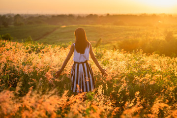 Trendy girl in stylish summer dress feeling free in the field with flowers in sunshine.