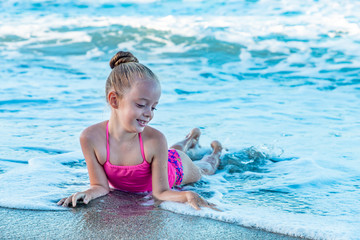 A playful smiling cute little girl in a colorful pink swimsuit lying in waves on the beach, ocean...