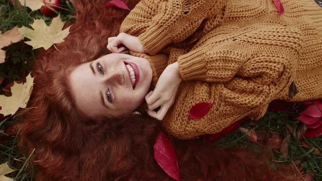 Young redhead, freckled beautiful happy smiling girl lays on grass and looks at camera. Top view