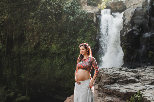 Young authentic pregnant woman near amazing cascade waterfall in Ubud, Indonesia. Beautiful morning sunlight, harmony with nature. Childbirth in Bali.