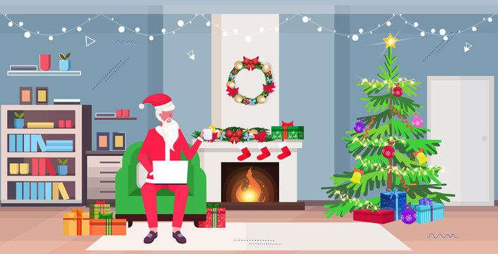 santa claus sitting in armchair using laptop and drinking tea modern living room with fireplace fir tree and gift boxes merry christmas new year holidays celebration concept horizontal full length