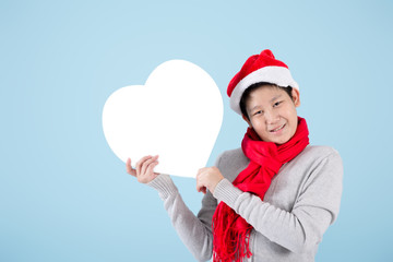 Happy Asian preteen boy wearing sweater and red scarf holding blank heart shape speech bubble on blue background.