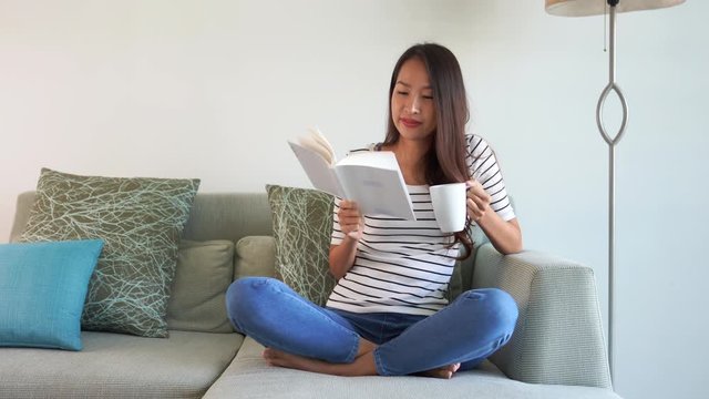 A young Asian woman relaxing in her jeans and a striped tee-shirt while holding a book in one hand and a cup of coffee in another.