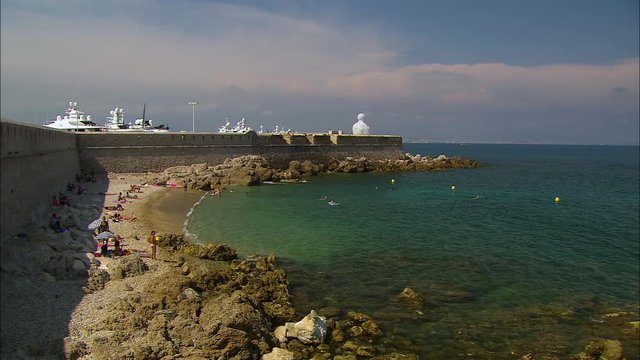 Wide low-angle still shot of tourist relaxing at a stony Antibes Beach near Vauban Port, French Riviera, France