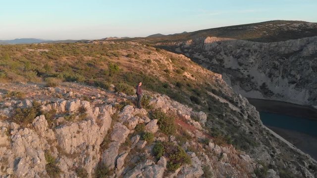Young caucasian stand over a cliff in zrmanja canyon, Croatia. Mountains landscape and wind turbines in the background. Drone cinematic golden hour