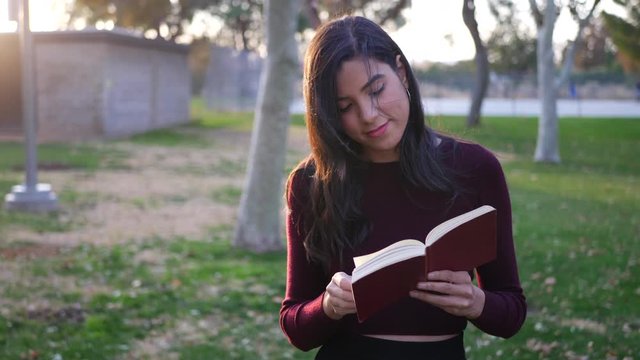 A young hispanic woman smiling and reading a story book or novel walking outdoors in the park at sunset SLOW MOTION.