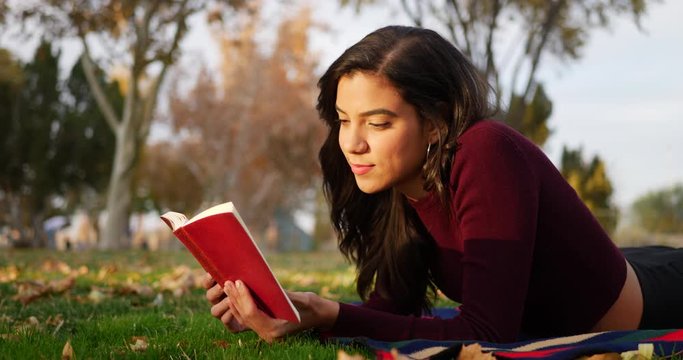 A hispanic woman college student reading a red book and smiling while laying down in the grass of a park at sunset.