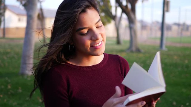 A Young Hispanic Woman Reading A Red Story Book Or Novel In The Park Smiling And Happy SLOW MOTION.