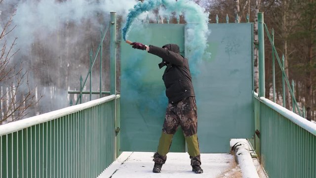 Man stay against closed steel doors on small bridge and wave hand with green smoke jet, full length shot. Winter outdoors, bare trees seen behind gate. Guy signal or mark position with colored smoke