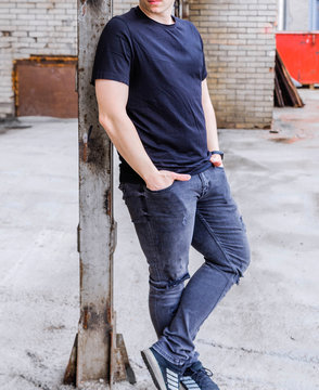 A young man poses for the camera in black jeans and a t-shirt. He holds his hands in his pockets.