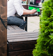 A young man in a white shirt and jeans sits at a table in a cafe on the street and looks at the phone