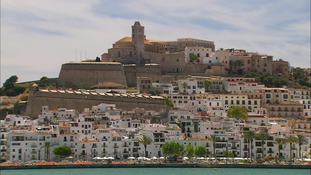 Low-angle still shot of an island hill with crowded white apartment blocks, and an ancient and historical building at the hilltop with long wind towers, island scene,  Ibiza Island, Spain