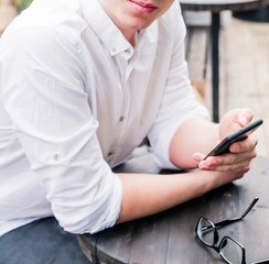 Fototapeta na wymiar A young man in a white shirt and jeans sits at a table in a cafe, holds a phone in his hands and looks at the camera