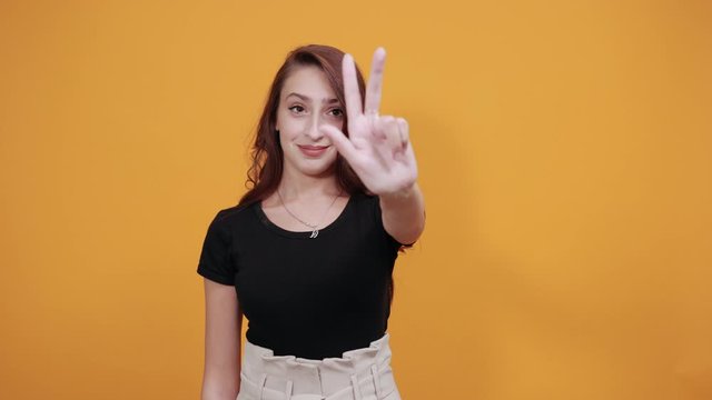 Attractive cheerful young woman in black shirt showing six fingers directly, smiling, laughting isolated on orange background in studio. People sincere emotions, health concept.