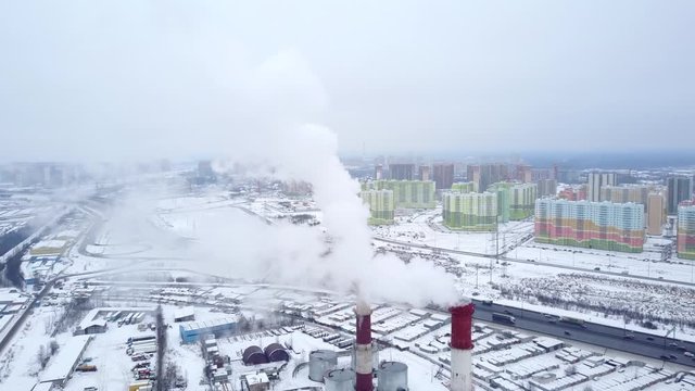 White clouds of smoke exhaust, two chimney stacks of heating plant, aerial orbiting shot. Residential blocks seen on background, industrial area and roads downwards. Typical cloudy weather at winter