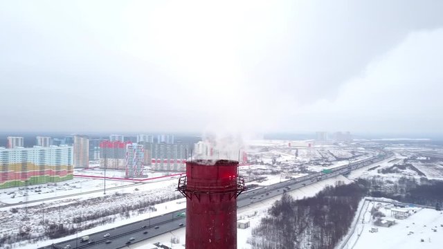 District heating plant chimney stack, smoke fly away from hole on top, orbiting aerial shot. Newly build colorful residential buildings seen on background, sleeping quarters at Saint Petersburg north