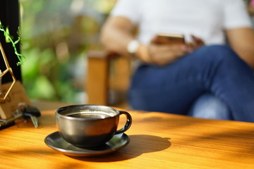 Hot coffee in a black ceramic mug placed on a wooden table in front of the woman who is sending a message via mobile phone.