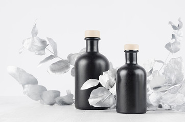 Elegant blank black bottles for cosmetic or home decor on soft light white wood table with silver leaves as template for advertising, design, branding identity.